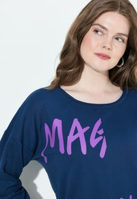 Maglietta blu a maniche lunghe con una grande stampa grafica viola. Il tessuto appare morbido con una vestibilità comoda e un collo a giro.