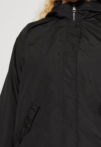 Schwarze leichte Jacke mit Kapuze, Reißverschluss und zwei seitlichen Taschen. Das Material wirkt glatt mit mattem Finish.