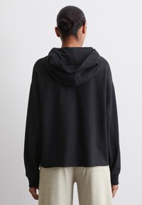 Zwarte hoodie met een relaxte pasvorm, lange mouwen en een zachte textuur, gecombineerd met lichtgrijze broek, gezien vanuit de achterkant.