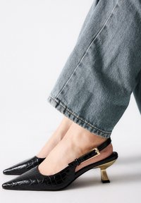 Zapatos slingback negros de piel con relieve de cocodrilo, con punta afilada y un tacón en bloque dorado, combinados con pantalones de mezclilla azul claro.