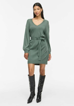 Femme portant une robe tricotée vert sauge ceinturée, manches longues, col en V festonné, associée à des bottes noires montantes à talons.