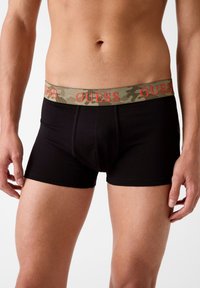 Boxer shorts neri con una cintura a motivo camouflage che presenta la scritta "GUESS" in testo arancione. Il materiale sembra morbido e ha un design aderente.