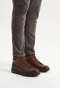 Brązowe skórzane buty trekkingowe z teksturowanym wykończeniem, okrągłym noskiem, metalowymi oczkami i ciemnymi gumowymi podeszwami, zestawione z szarymi spodniami z korduroju.