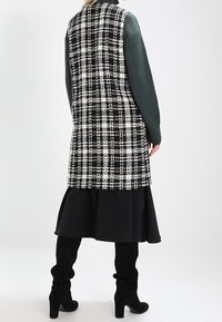 Długa czarno-biała kratowana kamizelka z teksturowanym tyłem, zielonymi prążkowanymi rękawami i czarną spódnicą. Noszona z czarnymi kozakami do kolan.