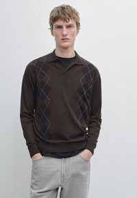 Maglione marrone lavorato a maglia con scollo a V e motivo a rombi in marrone scuro e nero. Maniche lunghe e orlo a coste, indossato sopra una camicia nera.