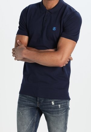 Mężczyzna w granatowej krótkim rękawie polo z emblematem, ze skrzyżowanymi ramionami, ubrany w niebieskie, postrzępione jeansy, na białym tle.