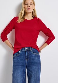 Röd ribbad långärmad topp med rund halsringning och knappade manschetter, kombinerad med blå denim byxor med vida ben och framfickor.