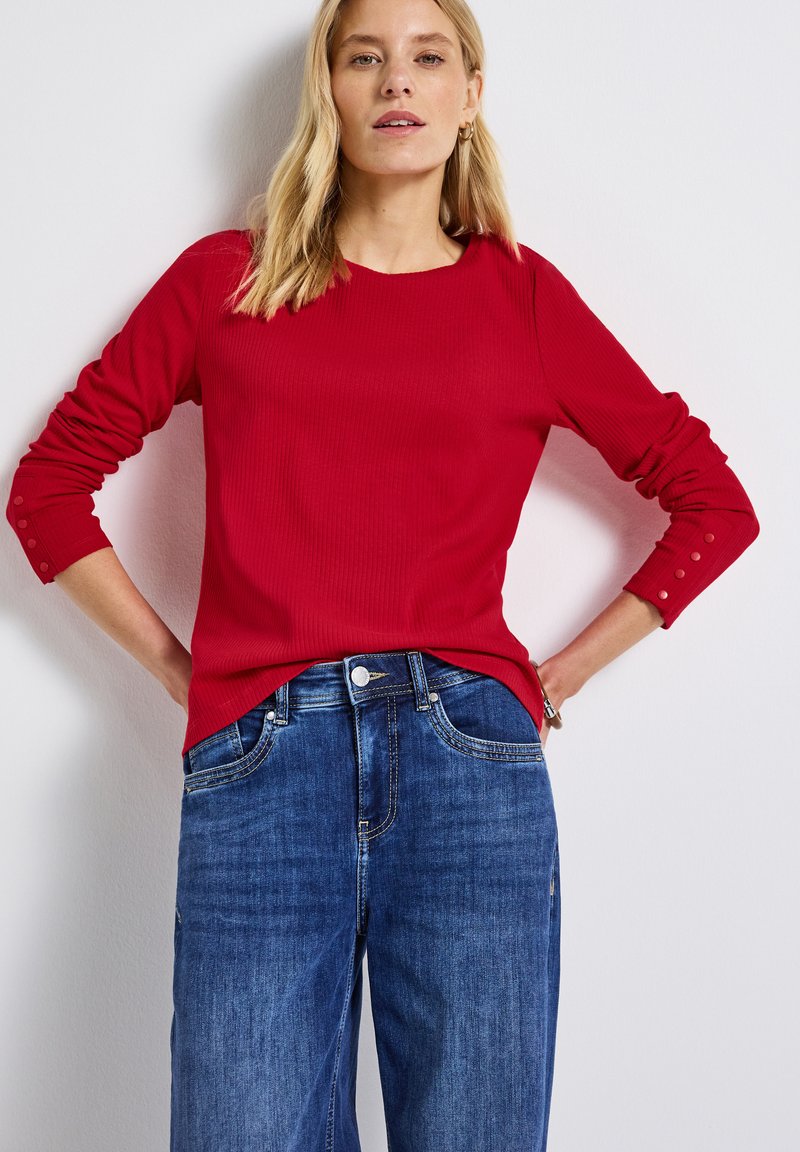 Röd ribbad långärmad topp med rund halsringning och knappade manschetter, kombinerad med blå denim byxor med vida ben och framfickor.