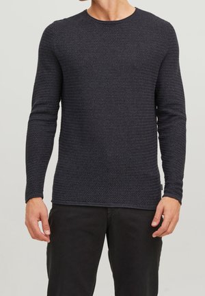 Man wearing black textured long-sleeve sweater and black pants, standing against a plain light background.