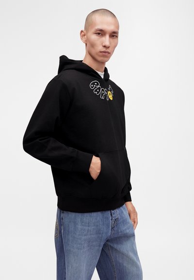 Homme portant un sweat à capuche noir avec un texte graphique et un smiley jaune, associé à un jean bleu, debout devant un fond blanc.
