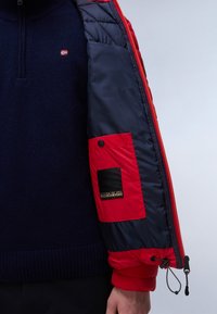 Syntetisk pufferjakke i rød og navy, med lynlås, ribbestrikket krave og en rød lomme med en sort logo-patch på ærmet.