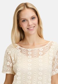 Kræmfarvet bluse i blonde med firkantet halsudskæring, korte ærmer og indviklede blomsterdesigns i stoffet.