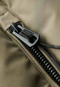 Olivgrön tyg med en svart dragkedja som har "SUPERDRY"-logotyp. Närbild visar dragkedjans tänder och texturerad dragpulldel för bättre grepp.