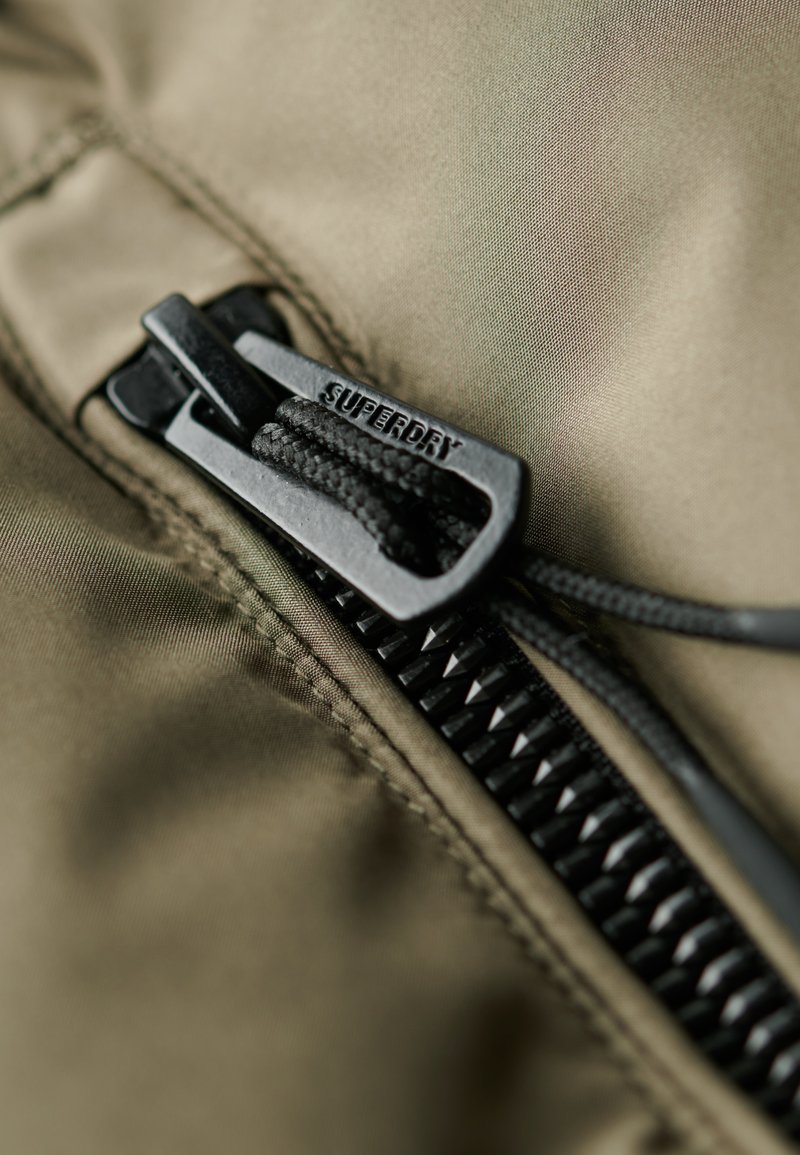 Olivgrüner Stoff mit einem schwarzen Reißverschluss, der die "SUPERDRY"-Markierung trägt. Nahaufnahme zeigt die Zähnchen des Reißverschlusses und den strukturierten Zugriegel für besseren Halt.