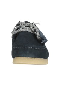 Navy mokasīnu kurpes ar noapaļotu purngalu un pelēkām un baltām rakstainām auklām, ar redzamu šuvju un gaišu zoli.