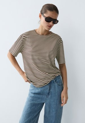 Femme portant des lunettes de soleil noires, un t-shirt à manches courtes rayé marron et blanc, et un jean bleu ample, debout avec une main sur la hanche.