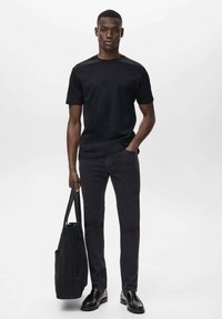 Camiseta negra de manga corta combinada con jeans oscuros y zapatos negros. La persona sostiene una bolsa de lona negra, de pie frente a un fondo claro.