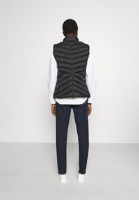 Černá prošívaná vesta s vodorovným prošitím, s vysokým límcem, nošená přes bílou košili, v kombinaci s tmavými džínami a bílými botami.