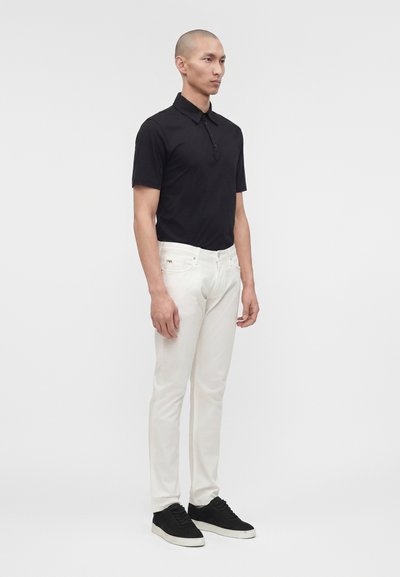Άνδρας που στέκεται, φοράει μαύρο πόλο με κοντά μανίκια, λευκό στενό παντελόνι τζιν και μαύρα αθλητικά παπούτσια πάνω σε απλό ανοιχτό φόντο.