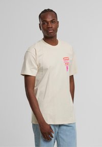 Beige bomullströja med korta ärmar, prydd med en rosa grafisk tryck på bröstet som inkluderar texten "KRÄV EN FÖRSLUTNING AV POLISBRUTALITET NU!"