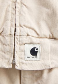 Fermeture éclair en tissu beige à texture côtelée, avec une étiquette logo Carhartt blanche à côté de la fermeture éclair.