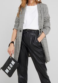 Femme portant un blazer gris à carreaux, une chemise blanche, un pantalon imitation cuir noir ceinturé, tenant un sac à main noirs MSGM dans sa main gauche.