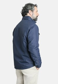 Chaqueta acolchada azul marino con cuello alto, que presenta un bolsillo en el pecho izquierdo y un detalle de puño abotonado, llevada sobre pantalones de color claro.
