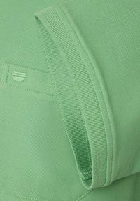 Miętowa dziana tkanina z prążkowanym kołnierzem i dołem. Posiada subtelny wzór i emblemat logo z przodu.