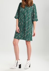 Vestido verde con un patrón floral, de corte suelto y mangas tres cuartos. Combinado con botas blancas hasta los tobillos con acentos negros y tachuelas.