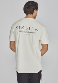 T-shirt en coton crème avec du texte imprimé noir au dos : "SIKSILK FollowtheMovement" avec des phrases supplémentaires en dessous.