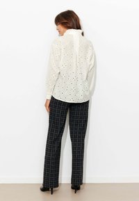 Blouse blanche à œillets avec découpes élaborées, col à volants, associée à un pantalon noir à carreaux taillé sur mesure, complété par des escarpins noirs à bouts pointus.