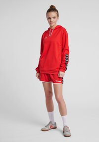 Rode hoodie met witte trekkoorden, gecombineerd met rode shorts met een logo. Zwart-witte chevronaccenten op de mouwen. Grijze sportschoenen.