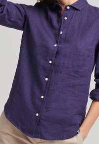 Kvinna iklädd en långärmad lila skjorta med vita knappar och beige byxor, med ena handen i fickan, framför en enfärgad bakgrund.