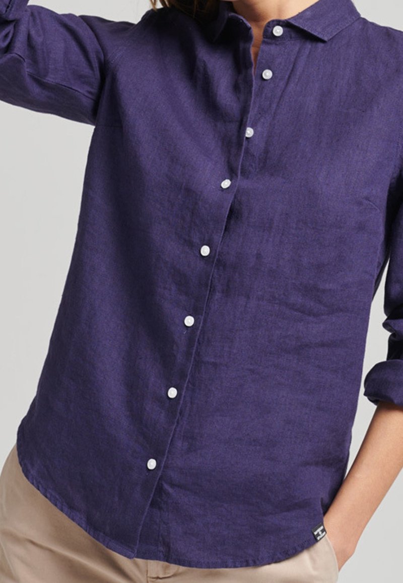 Kvinna iklädd en långärmad lila skjorta med vita knappar och beige byxor, med ena handen i fickan, framför en enfärgad bakgrund.