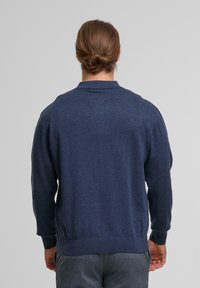 Mann mit braunen Haaren, die zu einem Dutt gebunden sind, trägt einen navyblauen Pullover und graue Hosen, steht mit dem Rücken zu einem schlichten Hintergrund.