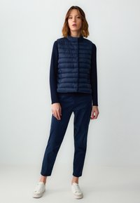 Marineblaue Steppweste mit gestepptem Muster, kombiniert mit navyblauen Slim-Fit-Hosen und weißen Sneakern. Die Weste verfügt über seitliche Reißverschlusstaschen.