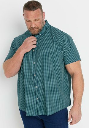 Mann, der den obersten Knopf eines kurzärmligen grün-navy karierten Hemdes schließt, trägt dunkelblaue Hose vor einfachem hellem Hintergrund.