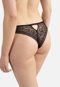 String noir en dentelle avec un motif floral, présentant un détail en forme de clé à l'arrière et une delicate finition le long des bords. Texture douce et couverture minimale.