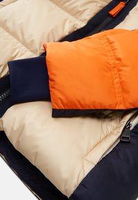 Puffer jacket with beige body, navy base, and orange cuff. Smooth fabric, quilted pattern, zipper detail, and contrasting colors.