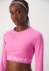 Rosa langermet crop top laget av elastisk materiale med rund halslinning og hvit Nike-logo. Har en kontrastliv med "PRO NIKE."