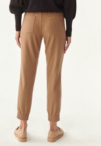 Brune, elastiske joggerbukser med en tætsiddende ankeldesign, der har to baglommer og en blød tekstur, båret med beige slip-on sko.