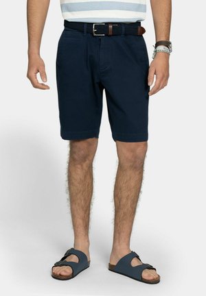 Mand iført navyshorts, et flettet bælte, en indstikket stribet skjorte og blå sandaler med spænder, stående med arme afslappet ned langs siden.