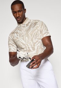 Beige Poloshirt mit weißen abstrakten Mustern, kurzen Ärmeln und Kragen; kombiniert mit weißen Hosen und einem Handschuh, der einen Golfschläger hält.