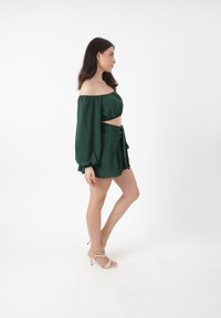 Kvinde i en grøn off-shoulder crop top og matchende nederdel, stående på siden med beige stropbehagelige sko mod en hvid baggrund.