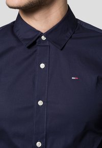 Navy blue button-up shirt in smooth fabric with a pointed collar, features clear plastic buttons and a small logo accent on the chest.