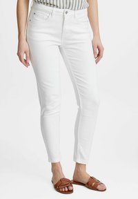 Bílé skinny džíny s hladkou texturou, s předními kapsami a jedním knoflíkem, doplněné hnědými plochými sandály.