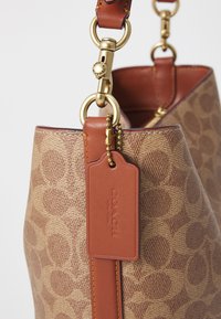 Brown canvas tote bag with a classic logo pattern, leather accents, and gold hardware. Includes a tag with embossed text and a sturdy strap.