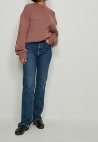 Różowy sweter z dzianiny o wysokim dekolcie, zestawiony z ciemnoniebieskimi rozkloszowanymi dżinsami oraz czarnymi chodakami z metalowymi, srebrnymi ćwiekami.