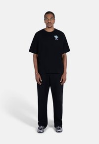 Svart oversized t-skjorte med et lite grønt og hvitt grafikkmotiv, kombinert med løse svarte bukser og grå sneakers. Enkel, minimalistisk design.