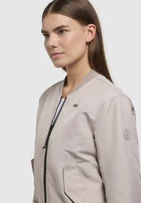 Helebeež bomber-jakk, millel on tõmbluku eesmine osa, sooniktagune kaelus ja küljetaskud. Varrel on ümmargune logo. Sile tekstuur.